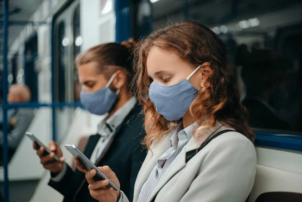 Two masked commuters sit on a train using their smartphones during their daily journey, illustrating the modern employee commute experience.