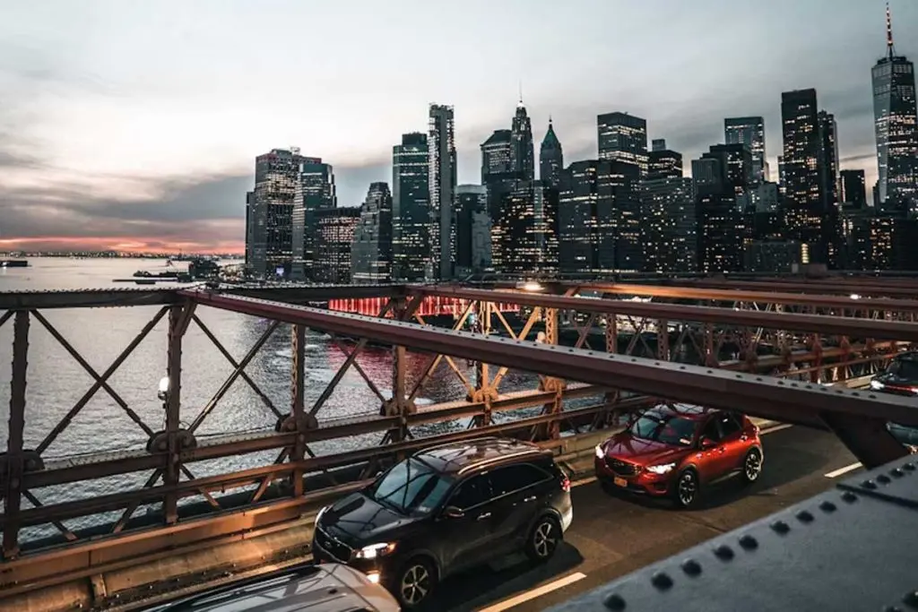 Cars crossing a steel-truss bridge at dusk with an illuminated skyline in the distance.