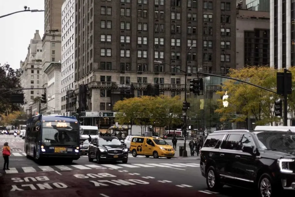 Transit bus, SUV, and taxi navigating a downtown intersection near a glass-front storefront.
