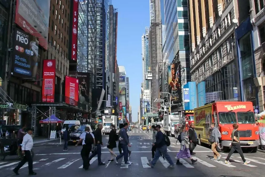 Pedestrians crossing a downtown intersection past parked delivery trucks and tall illuminated billboards.