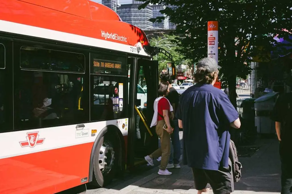 Hybrid-electric transit bus boarding passengers at a sunlit sidewalk stop lined with trees.