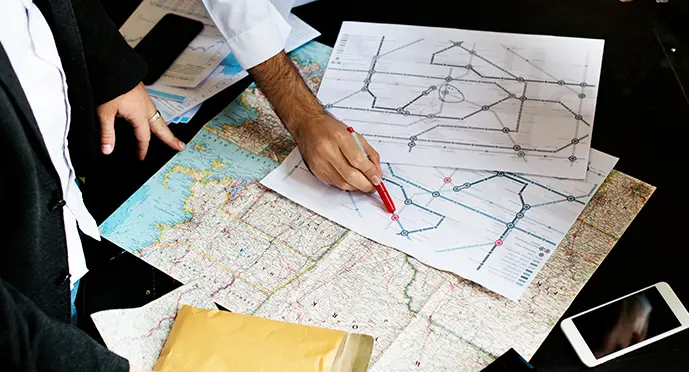 Two people reviewing transit route diagrams and marking nodes with a red pen over a topographic map.