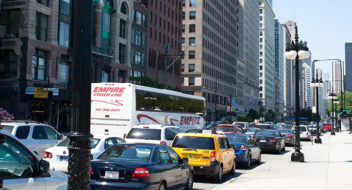 Congested city street with a charter bus, taxi, and sedans driving past historic brick buildings and skyscrapers.