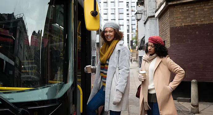 Two women in winter coats boarding a double-decker bus on a city sidewalk while holding coffee cups.