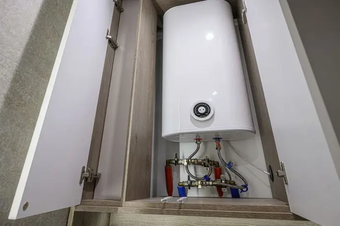A white hot water heater sits inside a cabinet with open doors, revealing pipes