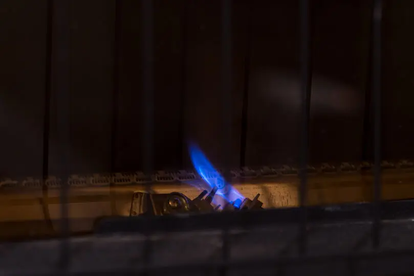 Small pilot flame for gas furnace heater. Constant blue flame for igniting main burners, behind safety grill