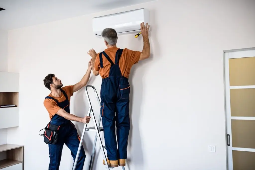Two workers in uniform, air conditioning masters using ladder while installing a new air conditioner in the apartment. Construction, maintenance and repair concept. Horizontal shot