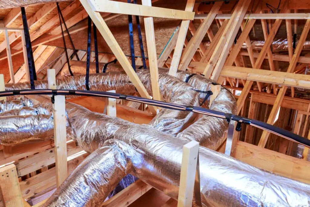 Workers install metal ductwork among wooden beams in home attic setting during renovation process.
