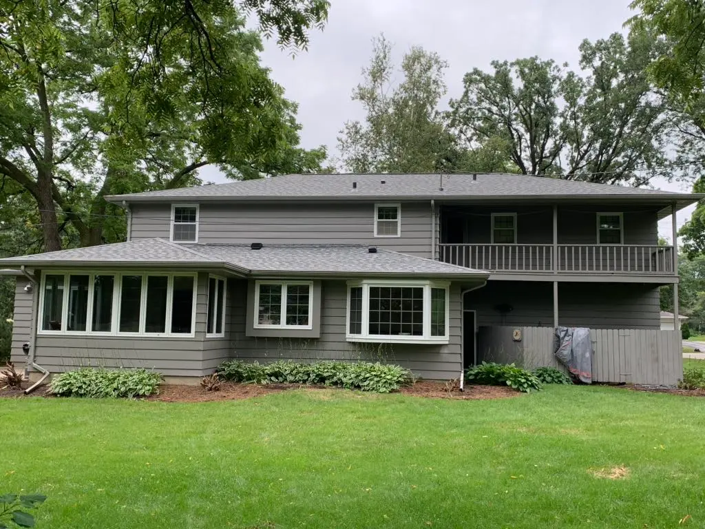 A gray two-story house with a sunroom, upper balcony, and well-maintained lawn, featuring quality roofing. Waunakee’s green trees and shrubs surround the home on a cloudy day.