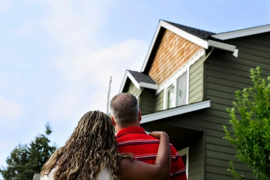 A couple stands close together, viewing a modern, green two-story house with a wooden accent under the roof. The sky is clear and blue, trees surround the home, and a solar installation hints at sustainability. The woman’s arm is around the man’s back, showing a sense of togetherness in Stoughton.