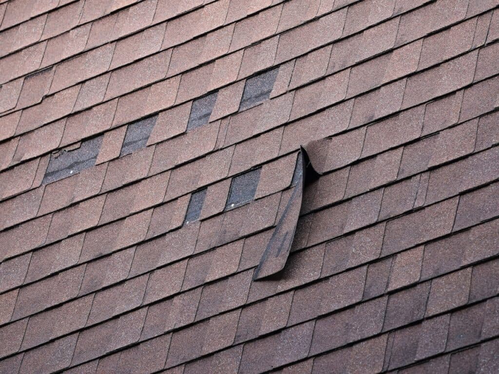 A section of a shingled roof with several misaligned shingles and one shingle partially detached, exposing the layer underneath. The shingles are a reddish-brown color, and the roof shows signs of wear.