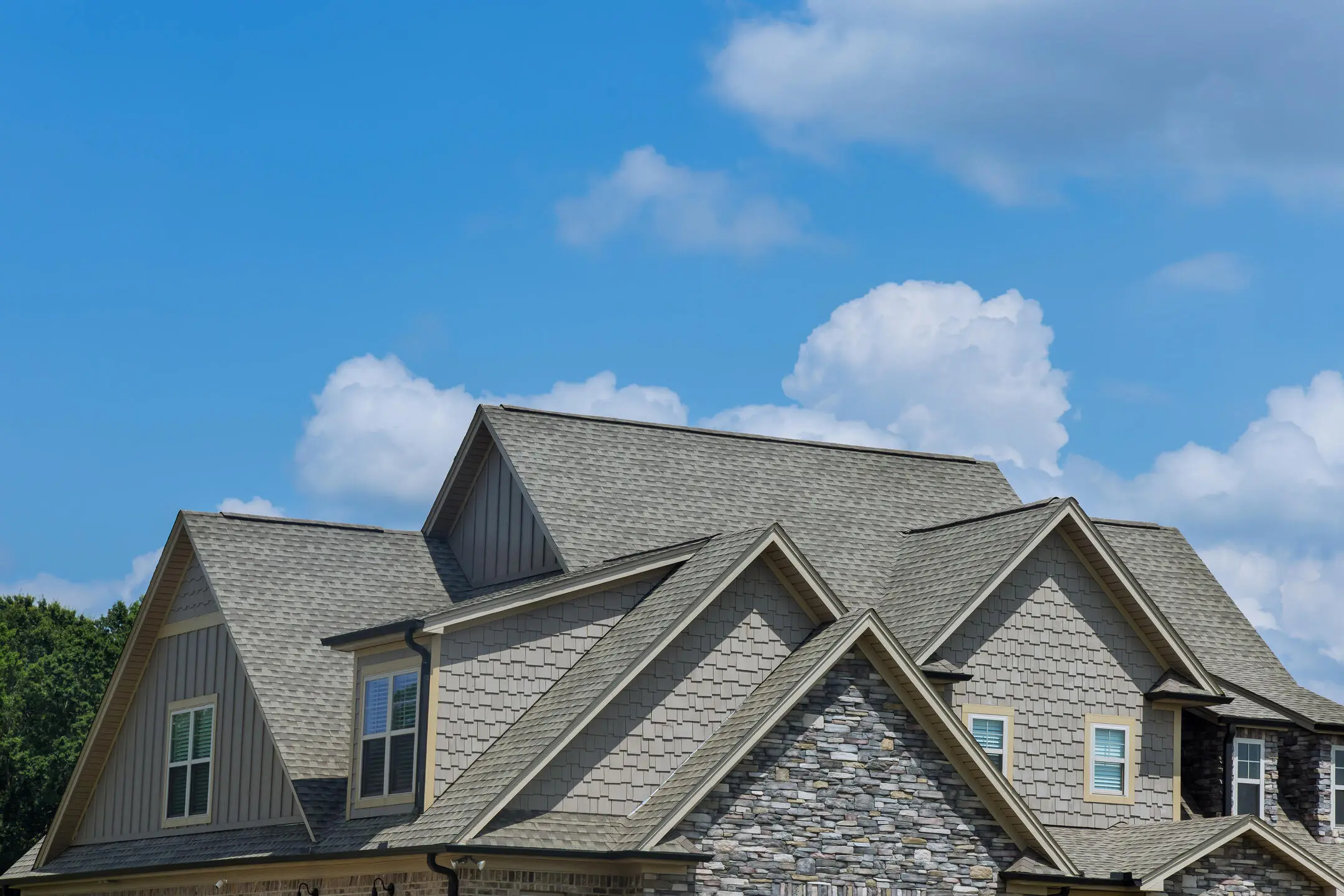 A house with intricate roof architecture, featuring multiple gables and a mix of shingles and stone siding. The roof is light gray, and the walls blend beige siding with stone accents. The sky is clear with a few fluffy clouds.