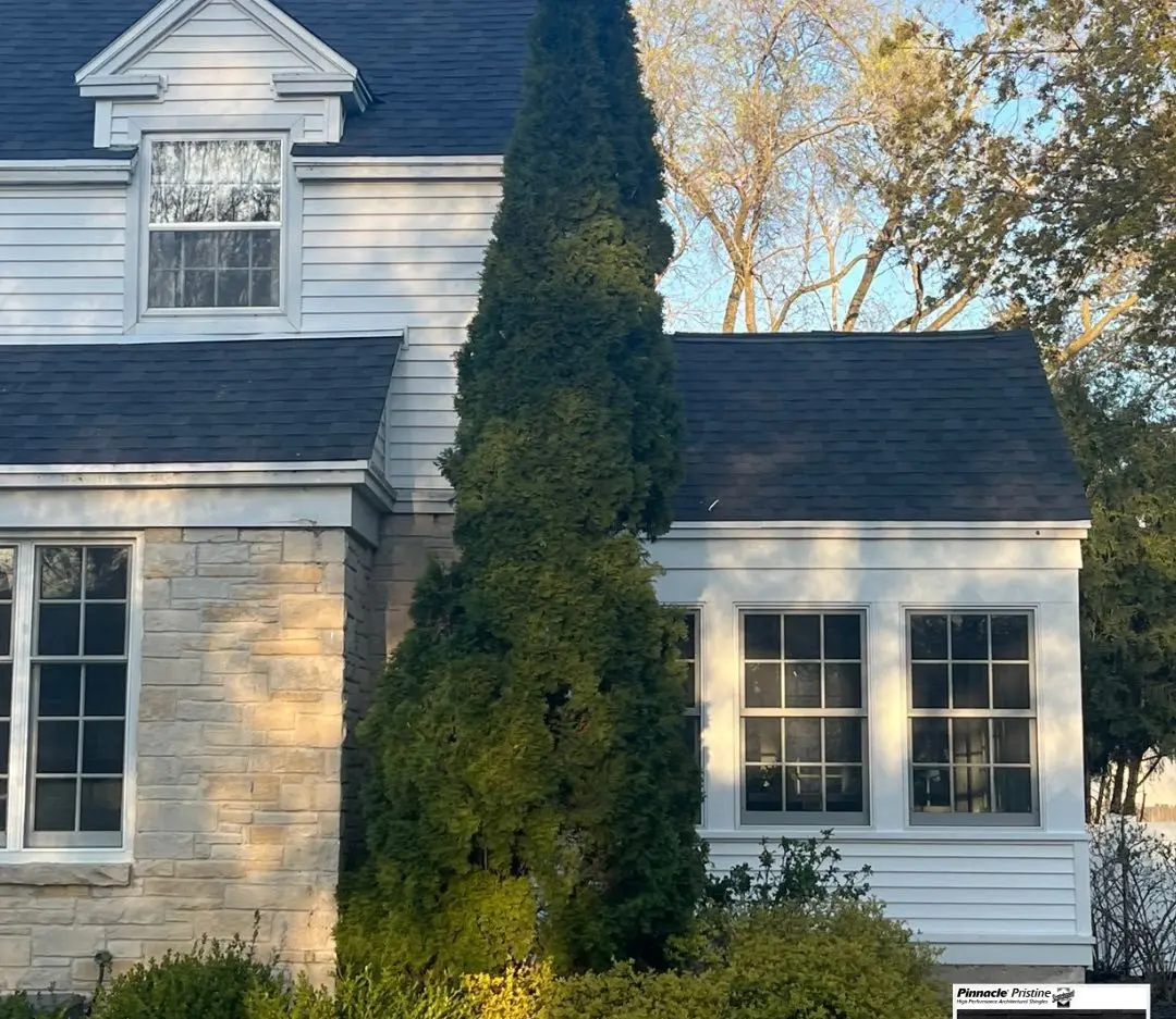 A tall, narrow evergreen tree stands in front of a white house with stone accents and multiple windows, its roofing Chenequa style bathed in soft sunlight.