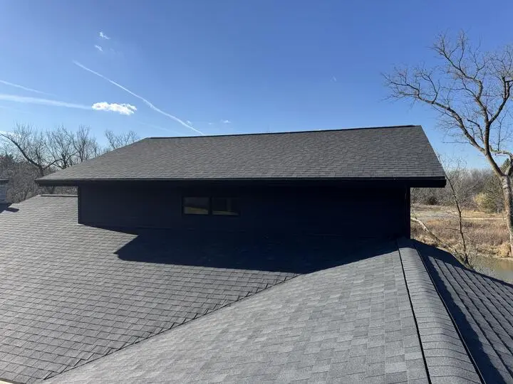 A house roof with dark gray shingles and a dormer featuring a small, rectangular window against a clear blue sky.