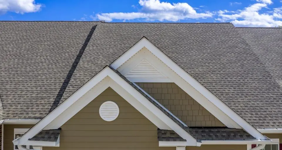 A close-up of a house roof featuring gray shingles and a gabled section with brown siding and a small round vent.