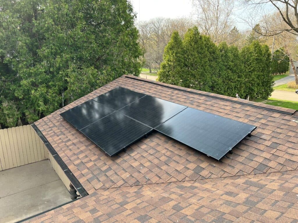 wisconsin solar incentives Three black solar panels are installed on the brown shingle roof of a house, surrounded by trees and a residential yard visible in the background.