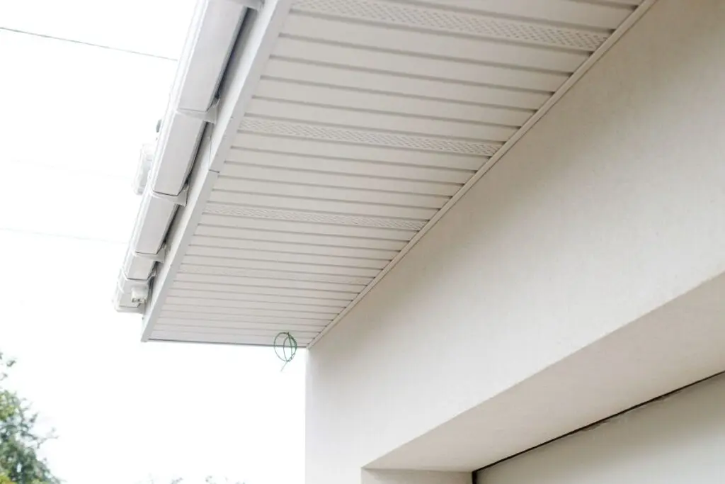 Close-up view of the underside of a house roof overhang with white soffit panels and an exterior wall, with a small green wire loop hanging from the soffit. Trees and power lines are visible in the background.