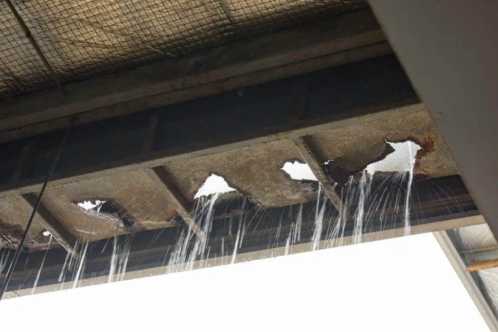 metal roof leak repair Water pours through large holes and rusted sections in a metal roof or ceiling, highlighting severe damage and corrosion against a bright sky—a clear sign that urgent metal roof leak repair is needed.