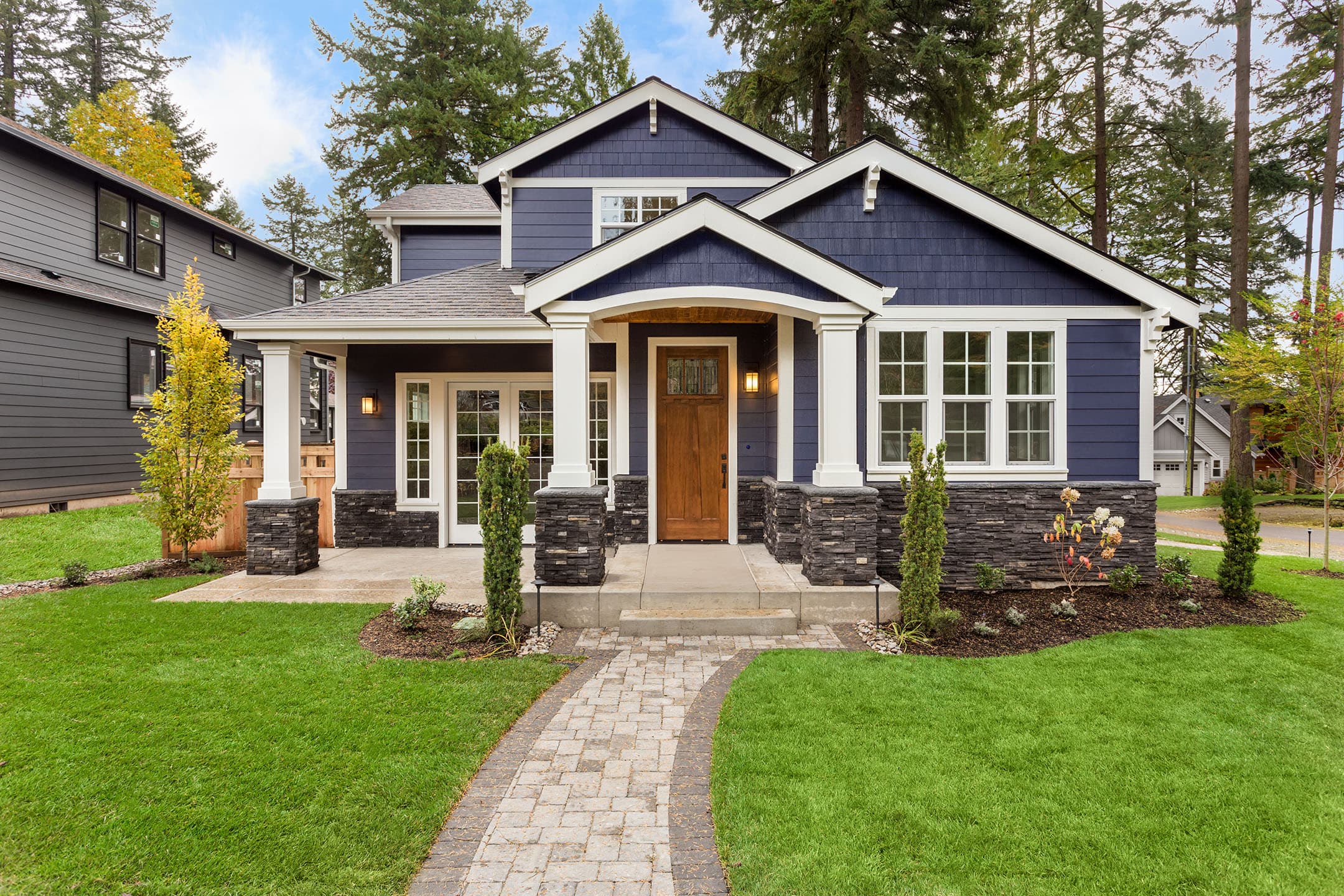 A charming modern house with blue siding, white trim, and a stone walkway. It features a wooden front door, multiple windows, and a well-kept lawn surrounded by tall trees on a sunny day.