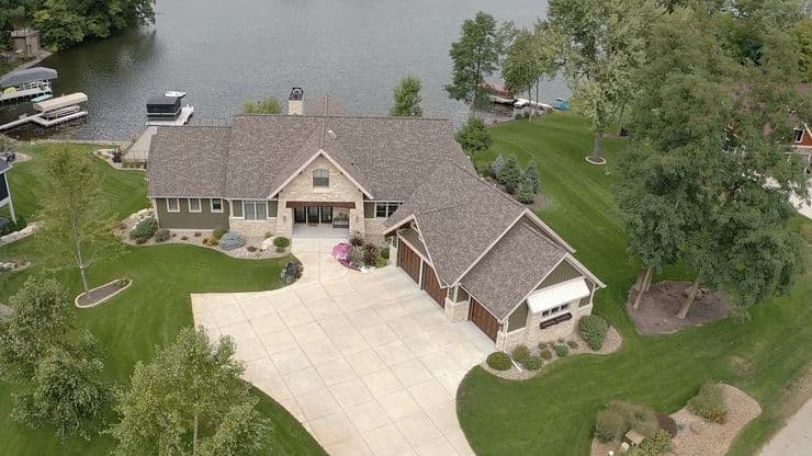 Aerial view of a large house with a gray roof and multiple gables, surrounded by green lawns and trees. The house features a spacious driveway leading to a three-car garage. In the background, there's a calm lake with a dock and boats.