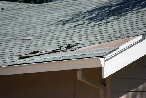 signs you need a new roof A house roof with damaged and missing shingles, exposing the underlying wood near the edge above the gutter.