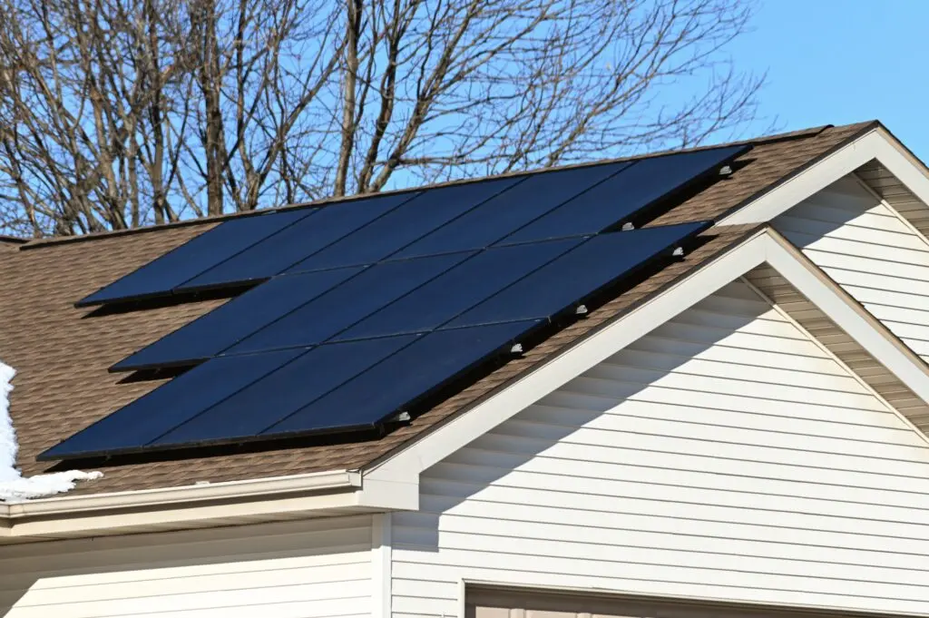 Solar panels installed on the roof of a suburban house with beige siding demonstrate how many solar panels to power a house; bare tree branches and a blue sky are visible in the background. how many solar panels to power a house
