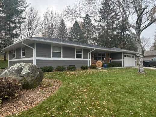 A gray ranch-style house with white trim, surrounded by trees and a well-kept lawn. There is a large rock in the foreground, scattered autumn leaves, and a driveway leading to an attached garage.