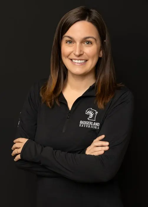A woman with straight brown hair and a friendly smile stands with arms crossed, wearing a black zip-up shirt with the Baggerland Exteriors logo, against a plain black background.