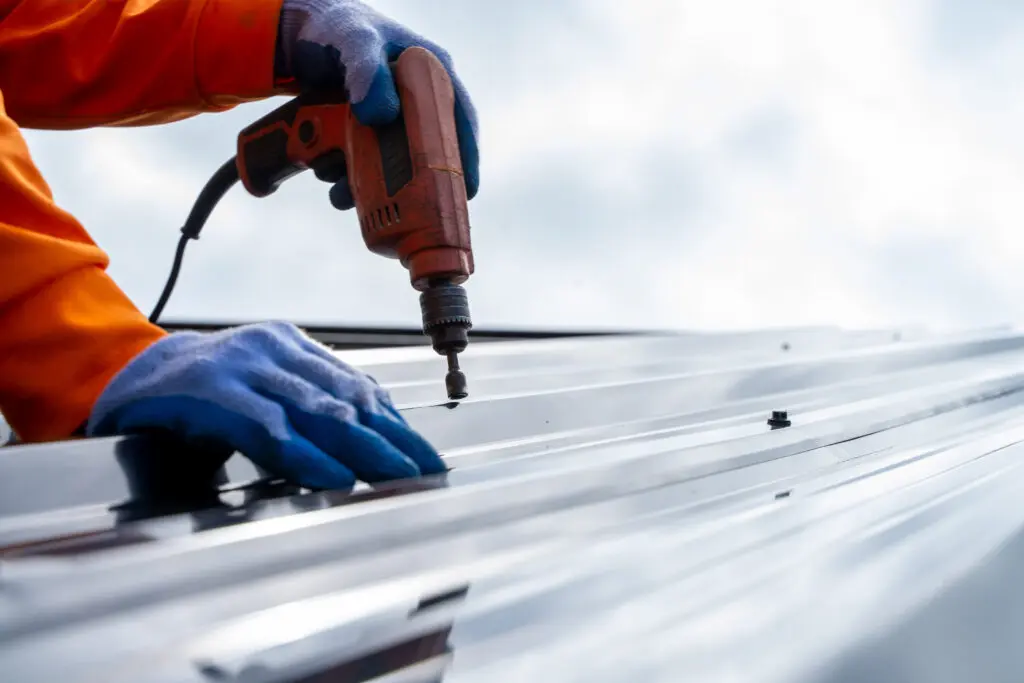 commercial roof repair services A person wearing gloves and an orange sleeve uses a power drill to fasten screws into a metal sheet, likely installing or repairing a roof. The sky is visible in the background.
