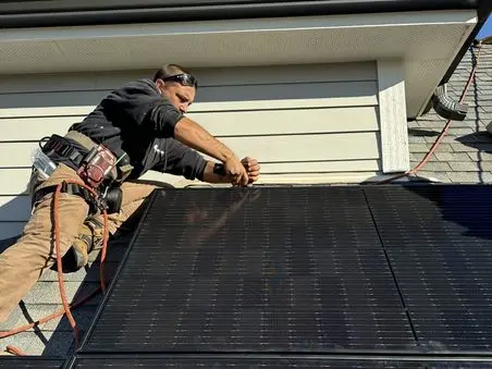A worker in safety gear installs a large black solar panel on a rooftop, using tools and harnesses, on a sunny day.