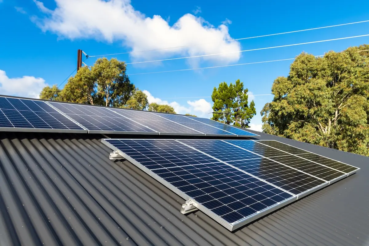 Solar panels are expertly installed on a dark corrugated roof under a clear blue sky, showcasing the precision of commercial roofing Madison. Tall trees stand in the background while power lines stretch across the scene, with panels angled perfectly to capture sunlight efficiently.