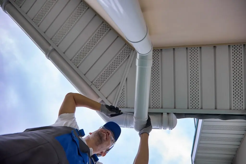 A worker in a blue cap and overalls installs or repairs a white rain gutter on the edge of a building roof, seen from a low angle against a partly cloudy sky.