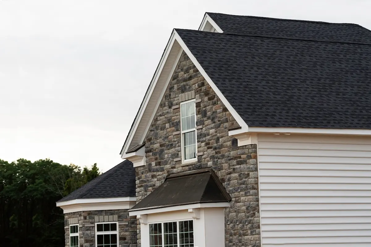 A close-up of a modern house exterior featuring gray stone and white siding, peaked roofs with dark shingles, and large windows. Trees are visible in the background under a cloudy sky.