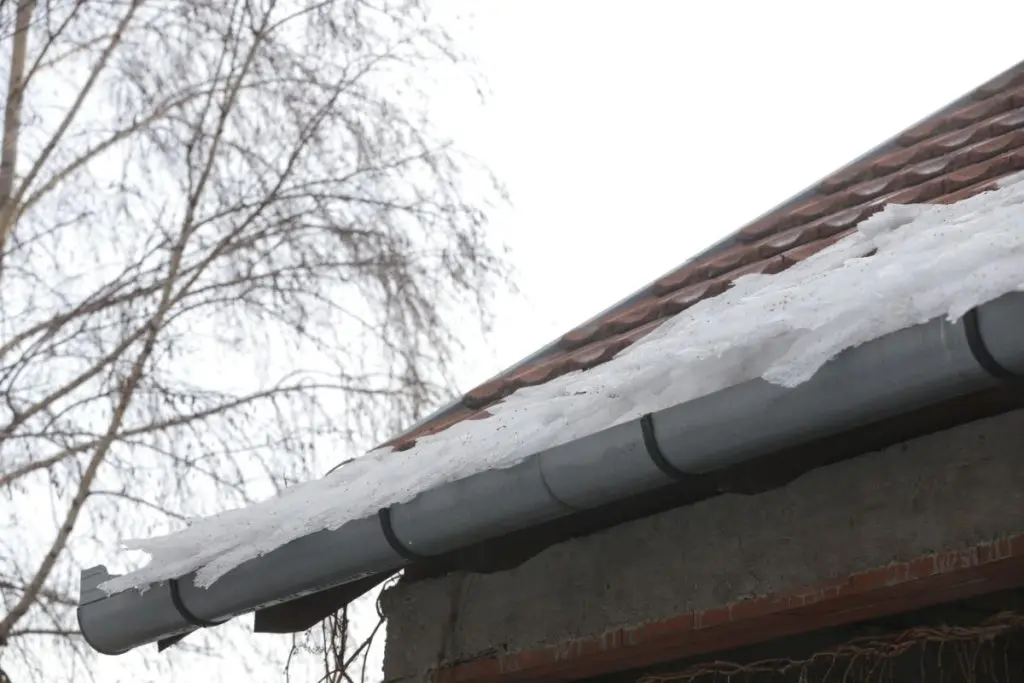 A close-up of a house roof with melting snow in the gutter. Bare tree branches are visible in the background against a cloudy sky.