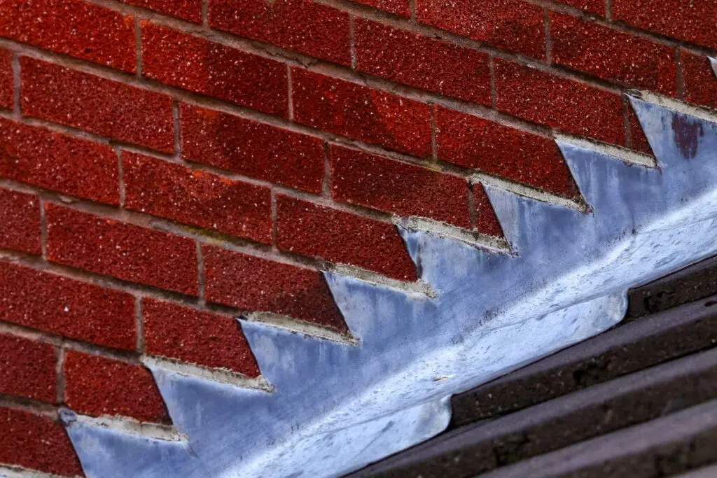A close-up view of red brickwork angled against a roof, with zigzag metal flashing where the bricks meet the roofline. The bricks are clean and evenly spaced, and the metal flashing is slightly weathered.