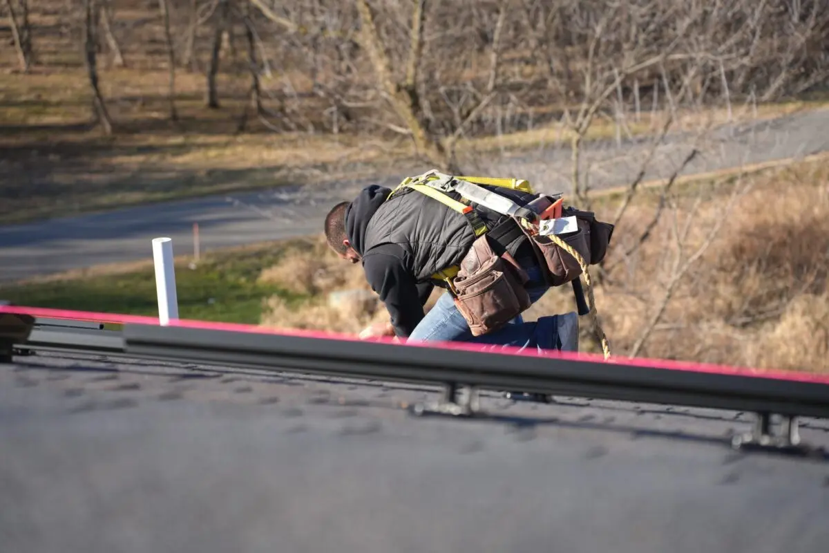roof maintenance checklist A worker wearing a safety harness bends over while working on a roof, with trees and a road in the blurred background.
