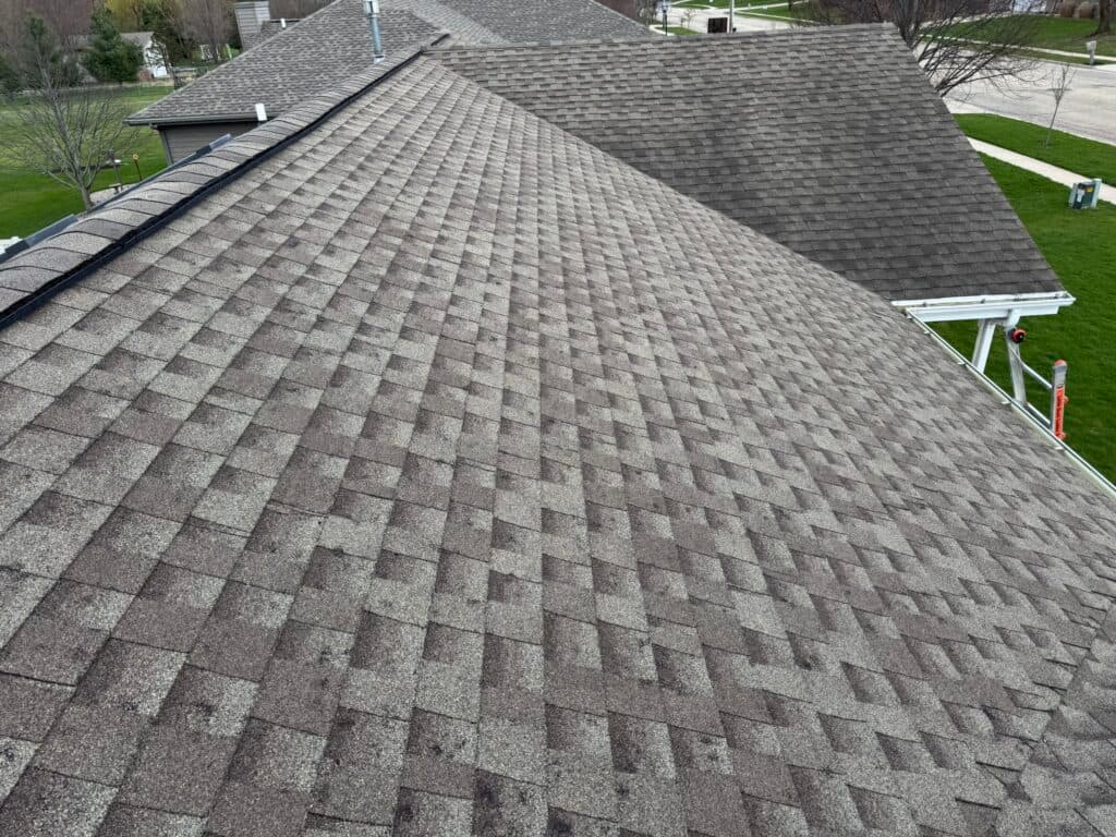 asphalt shingle roof replacement View of a sloped roof covered with brown asphalt shingles, with neighboring rooftops and a green lawn visible in the background. The shingles are arranged in an overlapping pattern.