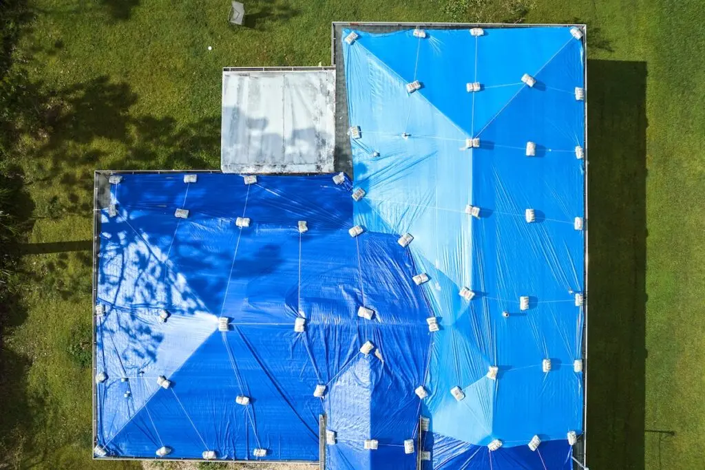 Aerial view of a flat-roofed building covered with blue tarps secured by weights. The surrounding area is grassy with patches of sunlight and shadows from nearby trees.