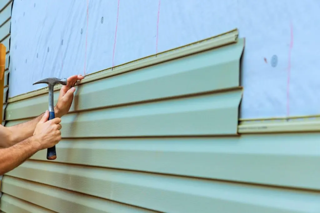 siding installation A person installs green vinyl siding on a house exterior, using a hammer to secure the material over a layer of building wrap.