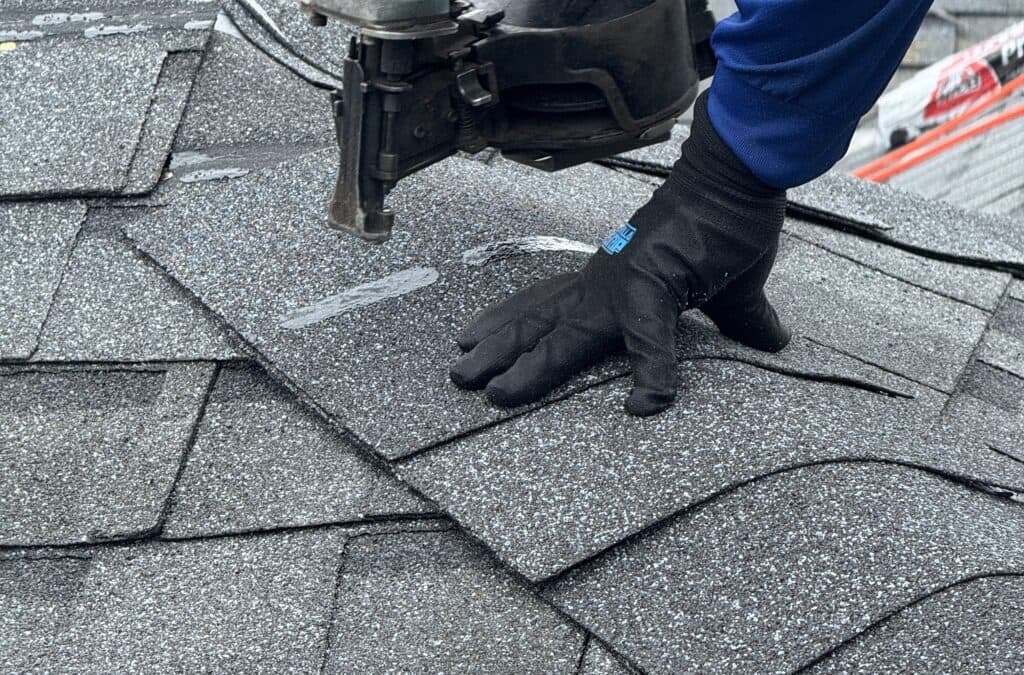 A person wearing a blue sleeve and black glove is using a nail gun to secure gray asphalt shingles on a roof.