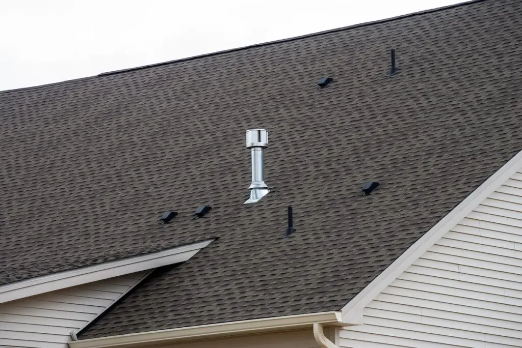 A steeply pitched house roof with brown shingles, a silver metal vent pipe, and multiple black snow guards installed in rows to prevent snow from sliding off. The house has light beige siding.