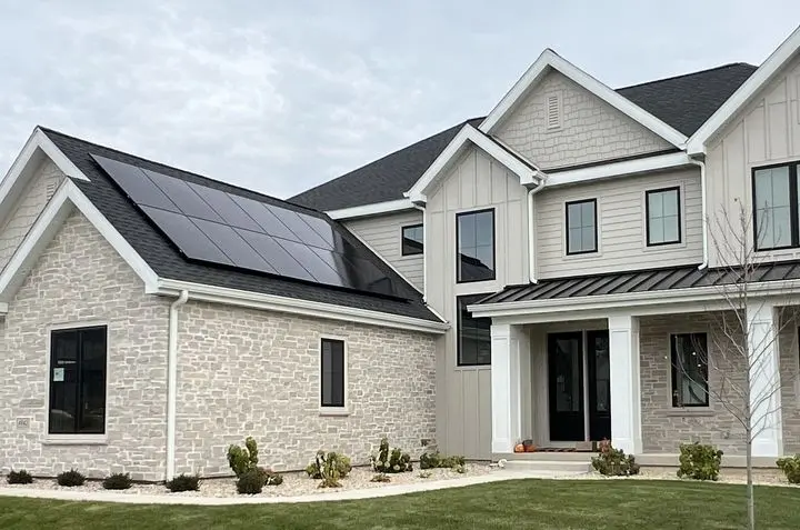 Modern two-story house with light-colored brick and siding, featuring solar panels on the roof above the garage. The front yard has neatly trimmed grass, small shrubs, and a covered entryway.