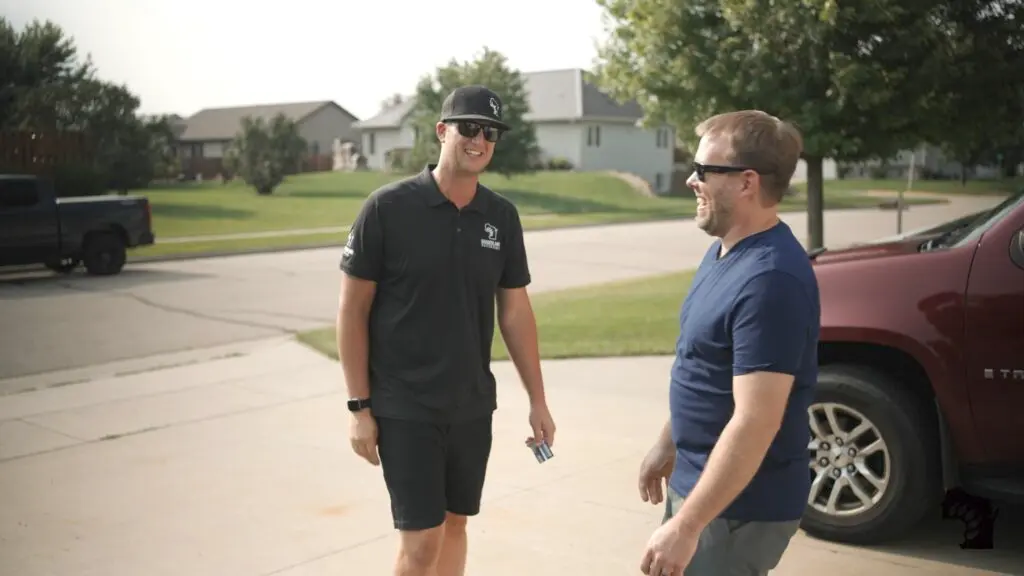 Two men are standing and smiling in a sunny suburban neighborhood. One wears a black polo, shorts, and cap, holding something in his hand. The other is in a blue shirt and sunglasses. A red SUV and houses are in the background.