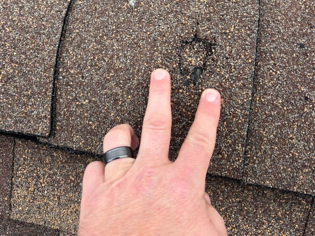 roof inspection services A hand with two fingers pointing at a small, circular indentation on brown asphalt roof shingles. The shingles appear weathered with some texture differences visible in the area.
