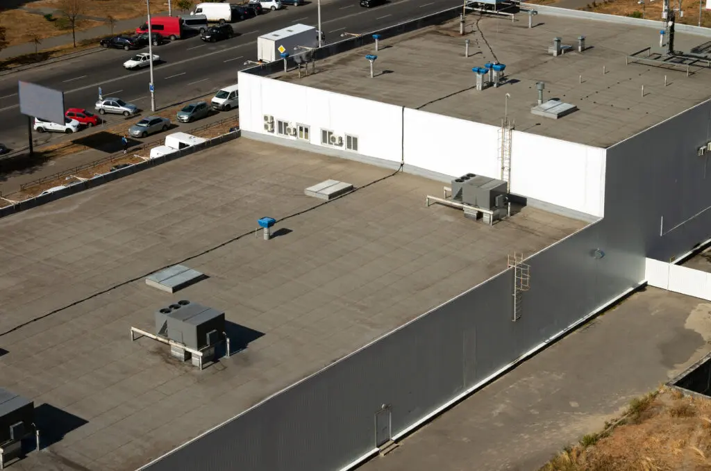 commercial flat roof repair A flat industrial building roof with HVAC units, vents, and ladders, next to a busy road with parked cars and grassy edges visible.