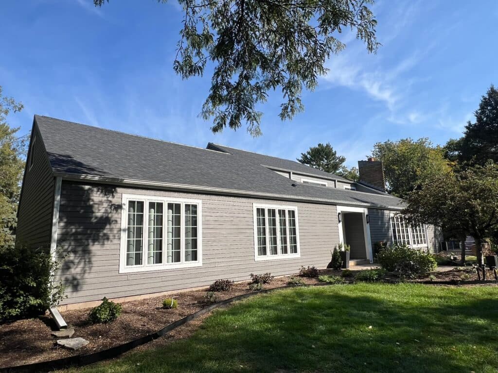 A single-story gray house with white-trimmed windows sits under a blue sky, surrounded by trees and a well-maintained lawn and garden, showcasing quality roofing McFarland homeowners trust.