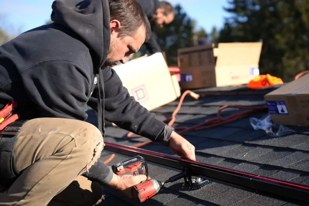 wisconsin solar incentives man in a black hoodie and tan pants installs equipment on a shingled roof using a power drill, with boxes and tools around him and another worker in the background.