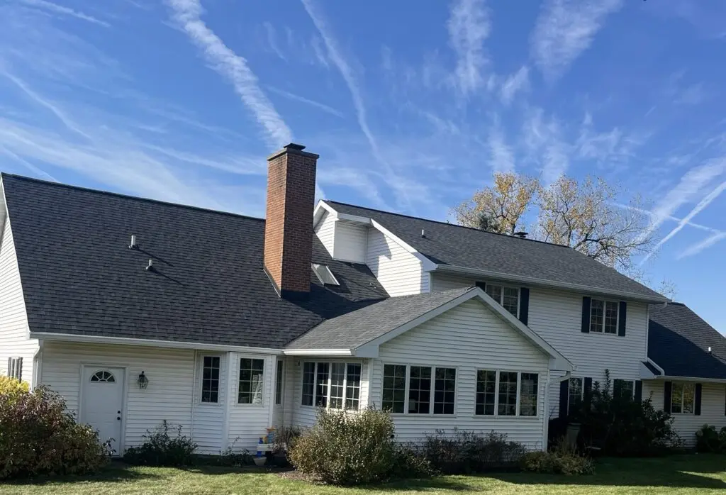 A two-story white house with a black roof by roofing Middleton and red brick chimney sits under a blue sky streaked with wispy clouds. Green bushes and grass surround the home, with some autumn trees in the background.