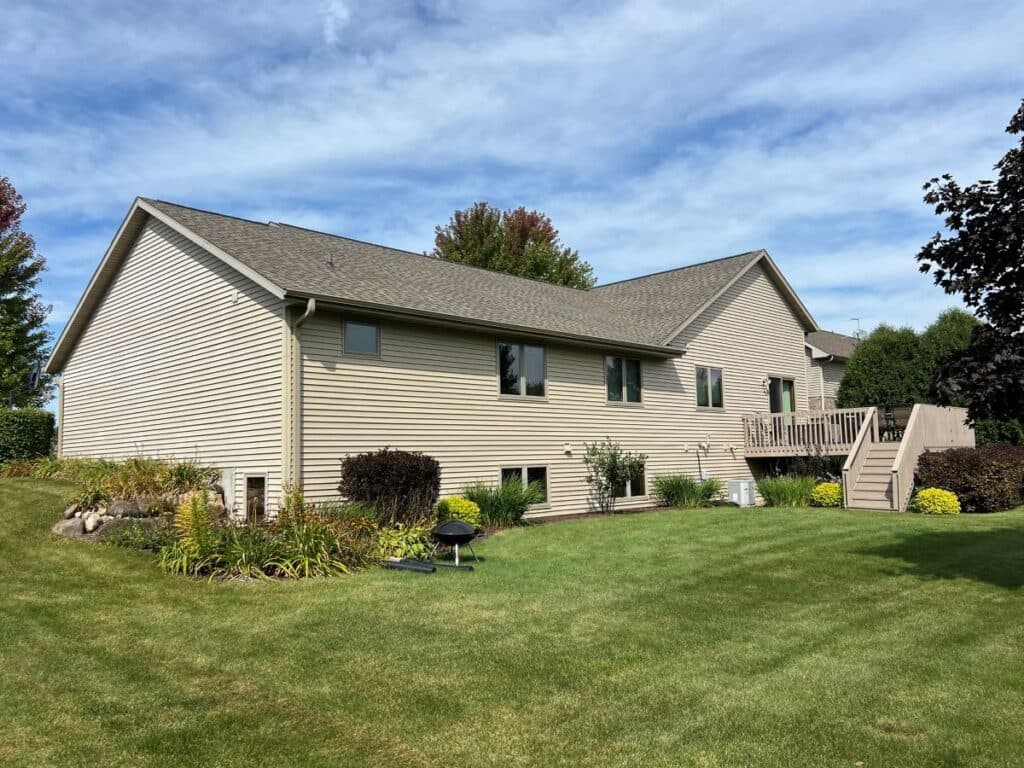 roof replacement timeline Single-story beige house with a sloped roof, several windows, and a wooden deck in the backyard. The yard is neatly landscaped with green grass, shrubs, and flowerbeds under a partly cloudy sky.