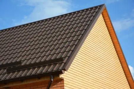 snow guards for metal roof Sloped roof with dark brown metal tiles on a yellow brick house, shown against a blue sky with scattered clouds.